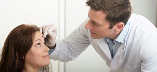 Das Bild steht für Botox Preise Wien. Es zeigt eine Szene im Dermacare Institut in Wien. Ein Arzt oder ein medizinischer Fachmann in einem weißen Kittel ist dabei, einer Patientin, die auf einem Behandlungsstuhl sitzt, eine Markierung auf der Stirn zu setzen. Dies ist eine Vorbereitung für eine Botox-Behandlung. Der Arzt trägt Einweghandschuhe und hält einen speziellen Stift, mit dem er präzise Markierungen auf der Stirn der Patientin setzt.