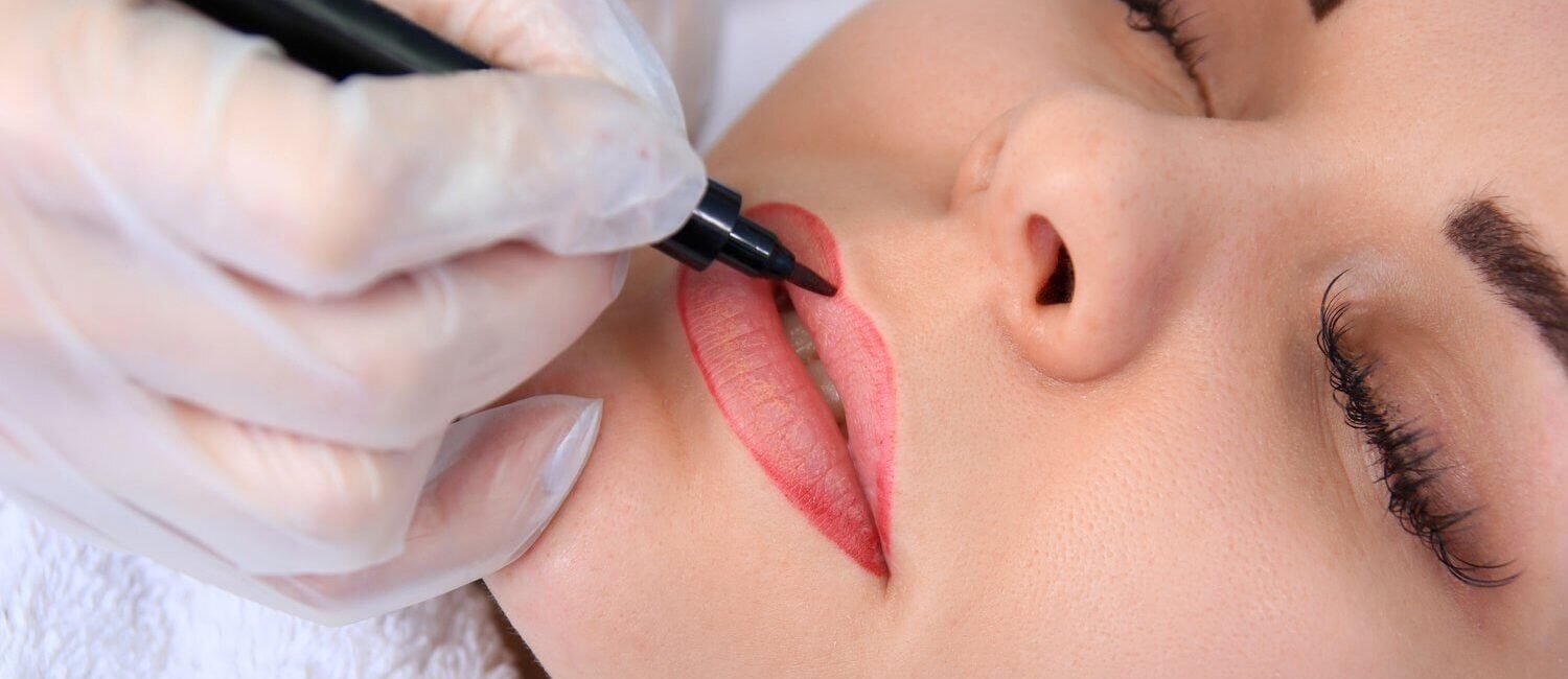 Das Bild zeigt eine Nahaufnahme einer Frau, die eine Permanent-Make-up-Behandlung für die Lippen erhält. Die Künstlerin, deren Hand mit einem Handschuh geschützt ist, trägt präzise rote Farbe auf die Lippenkontur der Kundin auf. Die Augen der Frau sind geschlossen, was darauf hindeutet, dass sie sich entspannt, während die Behandlung durchgeführt wird. Diese professionelle Permanent-Make-up-Behandlung wird von Dermacare Wien angeboten, wo höchste Präzision und individuelle Anpassung der Farbwahl für ein perfektes und langanhaltendes Ergebnis im Vordergrund stehen.