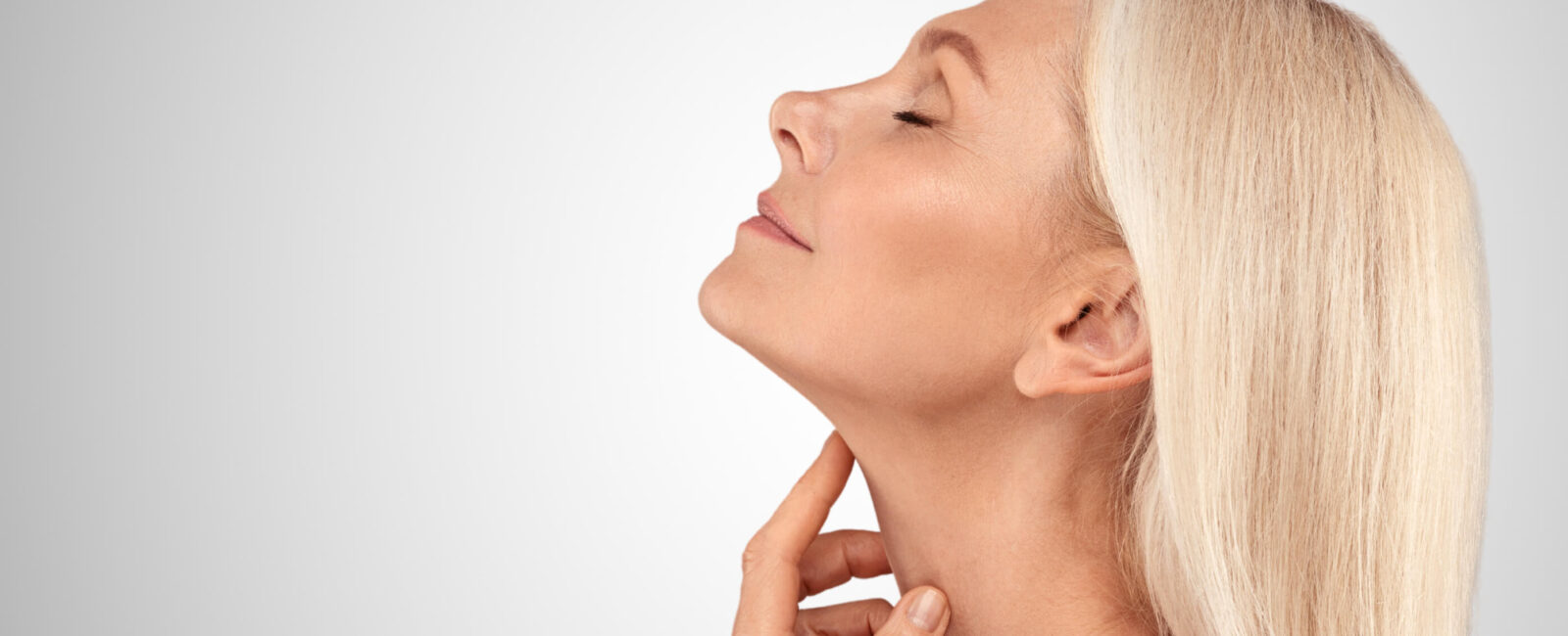 Das Bild zeigt eine blonde Frau, die ihr Kinn leicht anhebt und mit ihren Fingern sanft die Haut ihres Halses berührt. Sie wirkt entspannt und zufrieden, während sie ihren definierten Kinnbereich präsentiert. Ihr Gesichtsausdruck strahlt Zufriedenheit und Selbstbewusstsein aus, was die positiven Ergebnisse einer ästhetischen Behandlung wie die Entfernung eines Doppelkinns symbolisieren könnte. Der glatte Hals und das klar definierte Kinn weisen auf eine erfolgreiche nicht-invasive Behandlung hin, wie beispielsweise doppelkinn entfernen mit Radiofrequenz Microneedling oder andere Methoden, um das Doppelkinn ohne OP zu entfernen. Wenn Sie auch daran interessiert sind, Ihr Doppelkinn loszuwerden, könnte eine ästhetische Behandlung wie RF Microneedling die ideale Lösung sein!