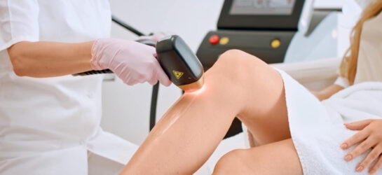 Eine professionelle Behandlung zur dauerhaften Haarentfernung bei Dermacare Wien. Eine Kosmetikerin in weißer Kleidung und rosa Handschuhen führt eine Laser Haarentfernung Wien an den Beinen einer Kundin durch. Die Kundin liegt entspannt auf einer Behandlungsliege und ist mit einem weißen Handtuch bedeckt. Die Spezialistin hält ein modernes Lasergerät, das mit einem roten Lichtimpuls auf die Haut trifft. Eine dünne Gelschicht ist aufgetragen, um die Haut während der Behandlung zu schützen. Neben der Laser Haarentfernung bietet Dermacare Wien auch die Nadelepilation als präzise Methode zur dauerhaften Haarentfernung an, um dauerhaft haarfrei zu sein.